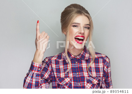 I have a idea. Satisfied beautiful girl with pink checkered shirt, collected updo hairstyle and makeup standing looking at camera with toothy smile, finger up. studio shot, isolated on gray background 89354611