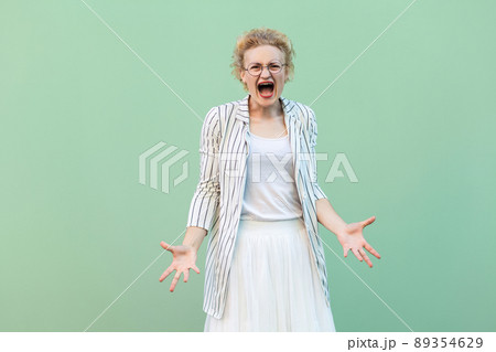 Portrait of angry young blonde woman in white shirt, skirt, and striped blouse with eyeglasses standing and screaming with raised arms. studio shot isolated on light green background 89354629