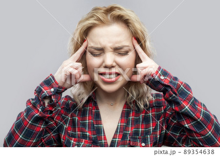 headache or confusion. Portrait of confused or sick blonde woman in casual red checkered shirt standing, touching temple, closed eyes and clenching teeth. studio shot, isolated on grey background. headache or confusion. Portrait of confused or sick blonde woman in casual red checkered shirt standing, touching temple, closed eyes and clenching teeth. studio shot, isolated on grey background. 89354638