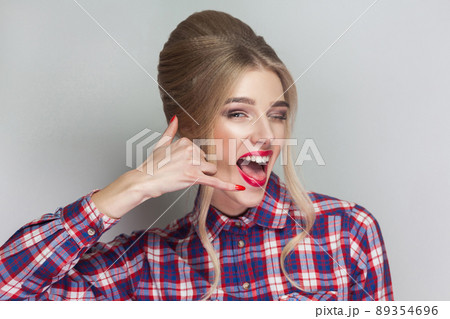 Call me! funny beautiful blonde girl in pink checkered shirt, collected updo hairstyle, red lips and makeup standing and looking at camera with calling gesture. indoor, isolated on gray background 89354696
