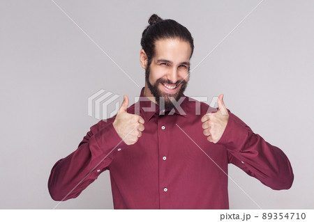 Portrait of handsome man with dark collected long hair and beard in red shirt standing, thumbs up and looking at camera satisfied, and toothy smile. indoor studio shot, isolated on gray background. Portrait of handsome man with dark collected long hair and beard in red shirt standing, thumbs up and looking at camera satisfied, and toothy smile. indoor studio shot, isolated on gray background. 89354710