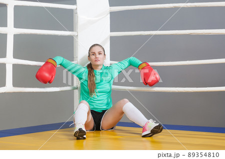 Portrait of confident beautiful young athlete woman with collected hair siting and leaning in the corner of boxing ring in red gloves and looking at camera. Indoor studio shot. on gray background. Portrait of confident beautiful young athlete woman with collected hair siting and leaning in the corner of boxing ring in red gloves and looking at camera. Indoor studio shot. on gray background. 89354810