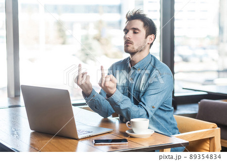 Fuck you! Side view portrait of angry upset bearded young big boss in blue jeans shirt are sitting in cafe, working and making video call on laptop, showing fucker and have conflict to the companion. 89354834