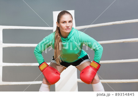 Angry boxing athletic young woman in green long sleeve poising with red boxing gloves and looking at camera with serious face. indoor studio shot on gray background. Angry boxing athletic young woman in green long sleeve poising with red boxing gloves and looking at camera with serious face. indoor studio shot on gray background. 89354845