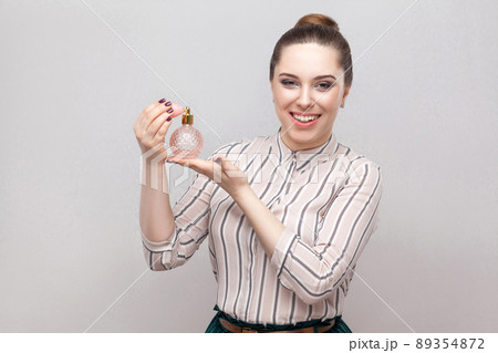 Woman using perfume. Smiling attractive woman spraying perfume enjoying aroma fragrance holding bottle of perfume isolated gray background. Indoor, studio sho, isolated on gray background, copy space Woman using perfume. Smiling attractive woman spraying perfume enjoying aroma fragrance holding bottle of perfume isolated gray background. Indoor, studio sho, isolated on gray background, copy space 89354872