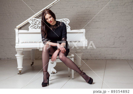 Sensual brunette woman in lace black classic dress sitting near white piano and looking at camera. indoor studio shot on white brick wall. Sensual brunette woman in lace black classic dress sitting near white piano and looking at camera. indoor studio shot on white brick wall. 89354892