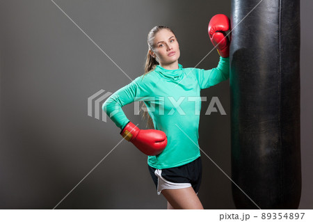 Portrait of sexy beautiful young athlete woman with collected hair standing, leaning against punching bag in boxing red gloves, looking at camera. Indoor studio shot, isolated on dark grey background. 89354897