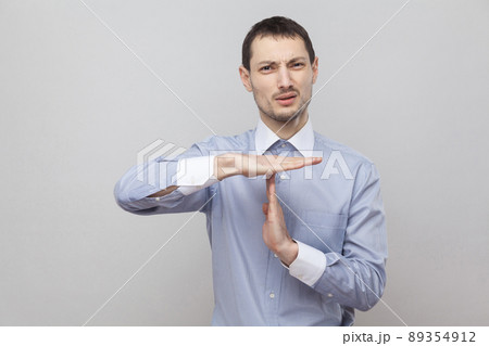 Please give me more time. Pleading handsome bristle businessman in classic blue shirt standing and looking at camera with timeout gesture. indoor studio shot, isolated on grey background copyspace. 89354912