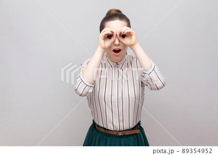 Portrait of funny beautiful young woman in striped shirt and green skirt with makeup and collected ban hairstyle, standing and looking with binoculars gesture. Studio shot, isolated on grey background Portrait of funny beautiful young woman in striped shirt and green skirt with makeup and collected ban hairstyle, standing and looking with binoculars gesture. Studio shot, isolated on grey background 89354952