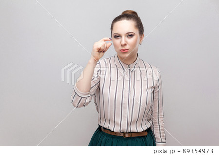 Give me a little bit more please. Portrait of hopeful beautiful young woman in striped shirt with makeup and collected ban hairstyle, standing and pleading. studio shot, isolated on grey background. 89354973