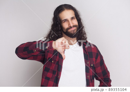 Portrait of unhappy handsome man with beard and black long curly hair in casual checkered red shirt standing looking at camera, showing dislike sign. indoor studio shot, isolated on grey background. Portrait of unhappy handsome man with beard and black long curly hair in casual checkered red shirt standing looking at camera, showing dislike sign. indoor studio shot, isolated on grey background. 89355104