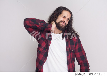 Portrait of sad handsome man with beard and black long curly hair in casual style, checkered red shirt standing and holding his painful neck with closed eyes. studio shot, isolated on grey background. Portrait of sad handsome man with beard and black long curly hair in casual style, checkered red shirt standing and holding his painful neck with closed eyes. studio shot, isolated on grey background. 89355129