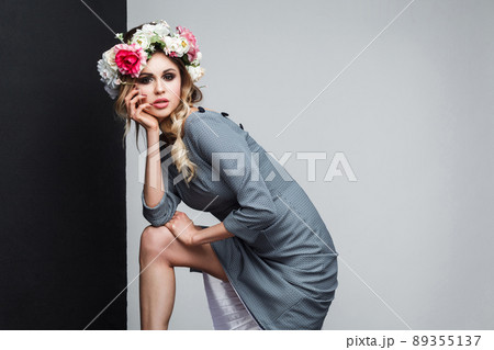 Portrait of beautiful sensual fashion model in grey dress with makeup and head flowers posing and looking at camera and touching her face. indoor studio shot, isolated on black and grey background. 89355137