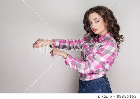 Time is gone. serious beautiful girl with pink checkered shirt, curly hairstyle and makeup standing showing time gesture and looking at camera. indoor studio shot, isolated on gray background. 89355165