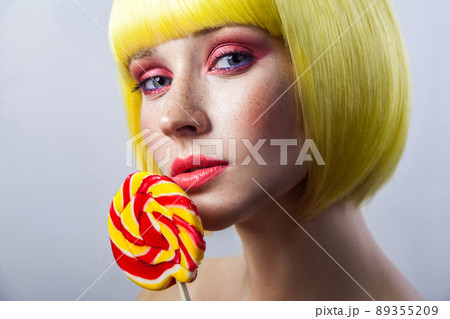 Beauty portrait of cute young female model with freckles, red makeup and yellow wig, holding colorful candy stick and looking at camera, serious face. indoor studio shot, isolated on gray background. 89355209