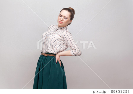Spine or kidney pain. Portrait of beautiful young woman in striped shirt and green skirt and collected ban hairstyle, standing and holding her painful back. studio shot, isolated on grey background. Spine or kidney pain. Portrait of beautiful young woman in striped shirt and green skirt and collected ban hairstyle, standing and holding her painful back. studio shot, isolated on grey background. 89355232