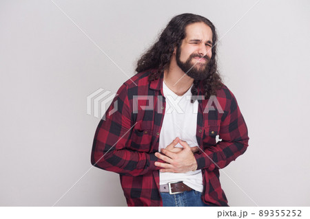 Portrait of sick handsome man with beard and black long curly hair in casual style, checkered red shirt standing holding his stomach with pain. indoor studio shot, isolated on grey background. 89355252