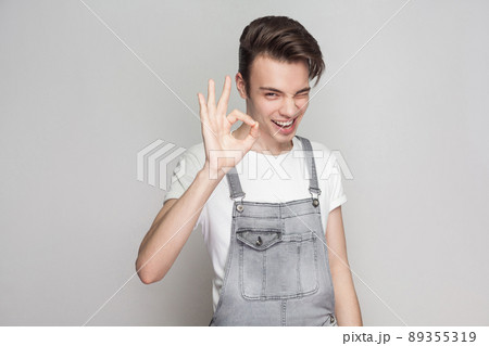 Happy young brunette man in casual style with t-shirt and denim overalls standing and looking at camera with toothy smile, winking and showing Ok sign. indoor studio shot, isolated on gray background Happy young brunette man in casual style with t-shirt and denim overalls standing and looking at camera with toothy smile, winking and showing Ok sign. indoor studio shot, isolated on gray background 89355319
