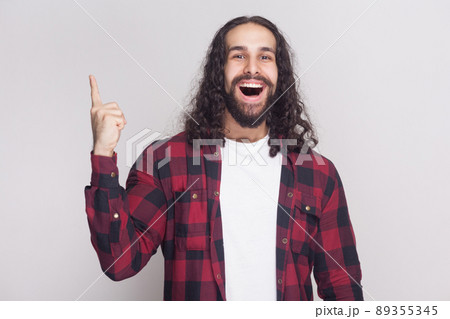 I have an Idea. happy amazed handsome man with beard and black long curly hair in casual checkered red shirt standing, looking at camera with finger up. indoor studio shot, isolated on grey background 89355345
