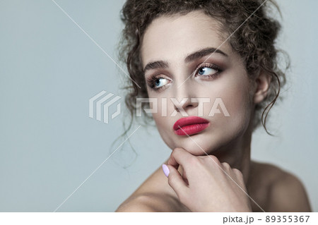 Beauty portrait of young beautiful fashion model with collected dark curly hair, nude makeup and red lips looking away serious with hand on chin. indoor studio shot. isolated on gray background. Beauty portrait of young beautiful fashion model with collected dark curly hair, nude makeup and red lips looking away serious with hand on chin. indoor studio shot. isolated on gray background. 89355367