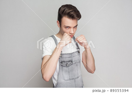 Portrait of serious young brunette man in casual style with t-shirt and denim overalls standing and looking at camera boxing fists and ready to attack. indoor studio shot, isolated on gray background. 89355394