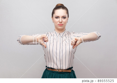 Unhappy beautiful young woman in striped shirt and green skirt with makeup and collected ban hairstyle, standing with thumbs down and looking at camera. indoor studio shot, isolated on grey background 89355525