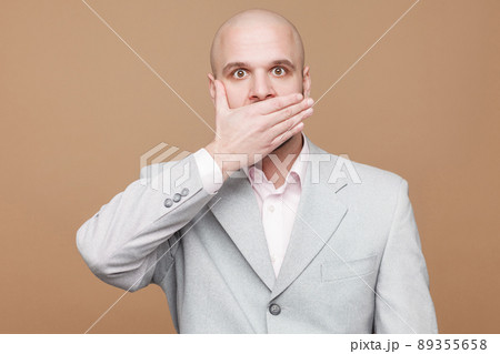 I'm will be quiet. Portrait of shocked handsome middle aged bald bearded businessman in classic light gray suit standing and covered his mouth. indoor studio shot, isolated on light brown background. 89355658