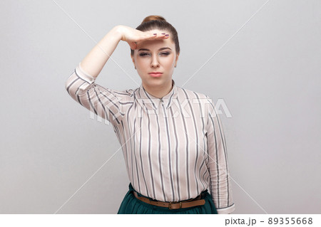Portrait of beautiful young woman in striped shirt and green skirt with makeup and collected ban hairstyle, standing and holding hand on forehead. indoor studio shot, isolated on grey background. 89355668
