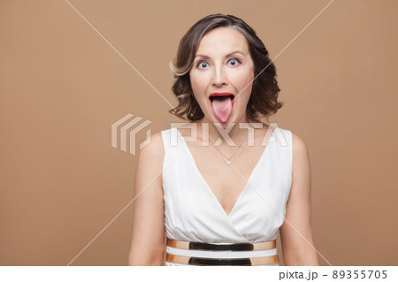 Crazy adult woman with big eyes tongue out . Emotional expressing woman in white dress, red lips and dark curly hairstyle. Studio shot, indoor, isolated on beige or light brown background 89355705