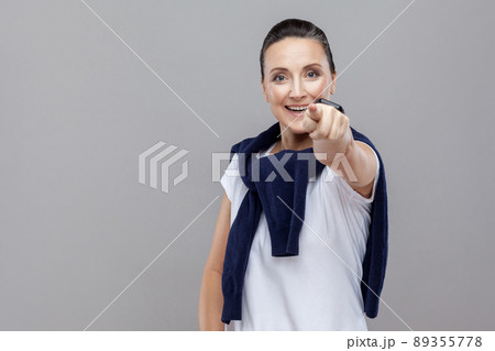 Hey you! Beautiful woman with blue jeans and sweater on her shoulders, pointing finger at camera, toothy smiling. Emotion and feelings Portrait of woman. Studio shot, isolated on gray background Hey you! Beautiful woman with blue jeans and sweater on her shoulders, pointing finger at camera, toothy smiling. Emotion and feelings Portrait of woman. Studio shot, isolated on gray background 89355778