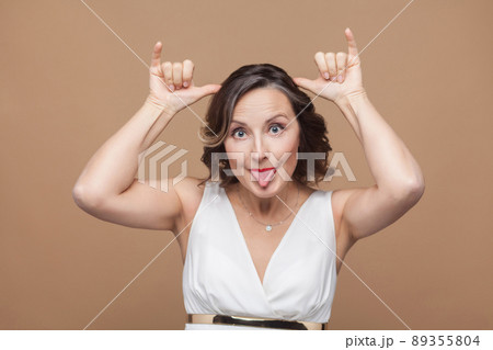 Funny adult woman making stupind mimicry at camera. Emotion and feeling concept. Studio shot, indoor, isolated on light brown background 89355804