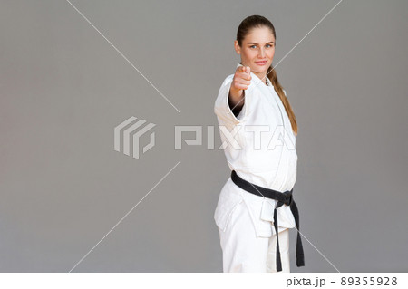 Confident athletic young woman in white kimono and black belt standing, pointing finger and looking at camera with smile. Japanese martial arts concept. Indoor studio shot, isolated on grey background Confident athletic young woman in white kimono and black belt standing, pointing finger and looking at camera with smile. Japanese martial arts concept. Indoor studio shot, isolated on grey background 89355928