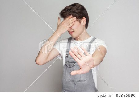 Portrait of scared or shy young brunette man in casual style with denim overalls standing closed eyes with hands, blocking and showing ban gesture. indoor studio shot, isolated on gray background. Portrait of scared or shy young brunette man in casual style with denim overalls standing closed eyes with hands, blocking and showing ban gesture. indoor studio shot, isolated on gray background. 89355990
