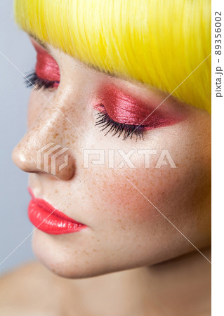 Closeup top view Beauty portrait of cute young calm female model with freckles, red makeup and yellow wig, closed eyes with serious face. indoor studio shot, isolated on gray background. Closeup top view Beauty portrait of cute young calm female model with freckles, red makeup and yellow wig, closed eyes with serious face. indoor studio shot, isolated on gray background. 89356002