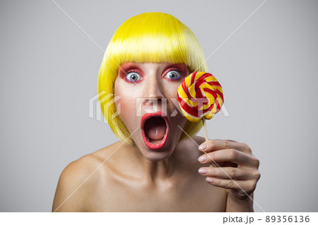 portrait of shocked cute young female model with freckles, red makeup, yellow wig holding colorful candy stick, looking at camera with surprised face. indoor studio shot, isolated on gray background. portrait of shocked cute young female model with freckles, red makeup, yellow wig holding colorful candy stick, looking at camera with surprised face. indoor studio shot, isolated on gray background. 89356136