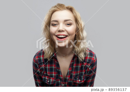 Portrait of happy beautiful blonde young woman in casual red checkered shirt standing, laughing and looking at camera with toothy smile. indoor studio shot, isolated on grey background. Portrait of happy beautiful blonde young woman in casual red checkered shirt standing, laughing and looking at camera with toothy smile. indoor studio shot, isolated on grey background. 89356137