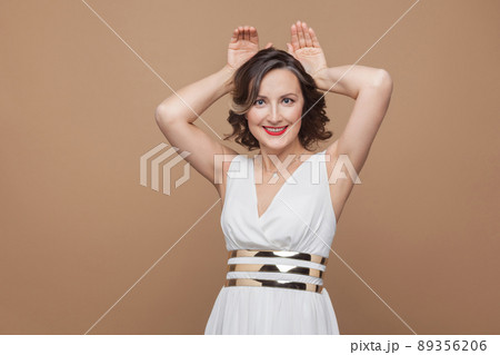 Funny middle aged woman showing rabbit ears and smiling. Emotional expressing woman in white dress, red lips and dark curly hairstyle. Studio shot, indoor, isolated on beige or light brown background Funny middle aged woman showing rabbit ears and smiling. Emotional expressing woman in white dress, red lips and dark curly hairstyle. Studio shot, indoor, isolated on beige or light brown background 89356206