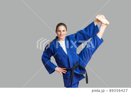 Beautiful happy athletic karate woman in blue kimono with black belt making vertical twine and looking at camera with toothy smile. Japanese martial arts concept. Indoor, studio shot, gray background Beautiful happy athletic karate woman in blue kimono with black belt making vertical twine and looking at camera with toothy smile. Japanese martial arts concept. Indoor, studio shot, gray background 89356427
