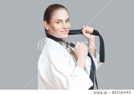 Side view profile of happy beautiful young athletic woman in white kimono with black belt wraps around the neck standing, holding belt and toothy smiling. Indoor studio shot, isolated, grey background Side view profile of happy beautiful young athletic woman in white kimono with black belt wraps around the neck standing, holding belt and toothy smiling. Indoor studio shot, isolated, grey background 89356449