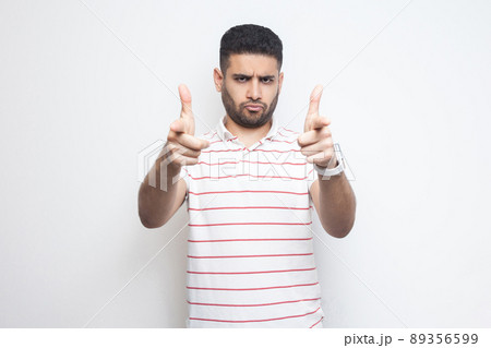 Hey you. Portrait of serious bossy handsome bearded young man in striped t-shirt standing, looking and pointing at camera. indoor studio shot, isolated on white background. Hey you. Portrait of serious bossy handsome bearded young man in striped t-shirt standing, looking and pointing at camera. indoor studio shot, isolated on white background. 89356599