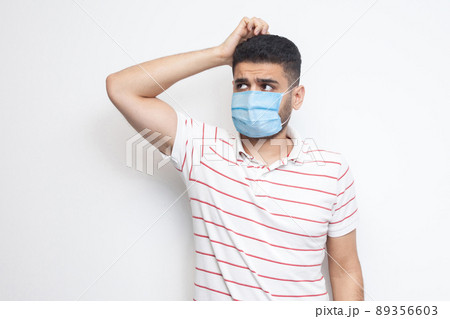 Portrait of query young man with surgical medical mask in striped t-shirt standing scratching his head, planing and thinking about something. indoor studio shot, isolated on white background. 89356603