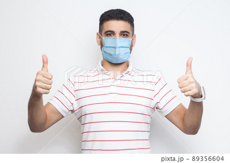I like this. I am safe. Portrait of satisfied happy young man with surgical medical mask in striped t-shirt standing, thumbs up and looking at camera. indoor studio shot, isolated on white background. I like this. I am safe. Portrait of satisfied happy young man with surgical medical mask in striped t-shirt standing, thumbs up and looking at camera. indoor studio shot, isolated on white background. 89356604