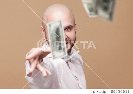 Portrait of happy handsome middle aged bald bearded businessman in white shirt standing, throw out money and looking at camera with proud rich face. studio shot, isolated on light brown background. 89356611