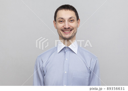 Portrait of happy satisfied handsome bristle businessman in classic light blue shirt standing and looking at camera with toothy smile. indoor studio shot, isolated on grey background copyspace. Portrait of happy satisfied handsome bristle businessman in classic light blue shirt standing and looking at camera with toothy smile. indoor studio shot, isolated on grey background copyspace. 89356631
