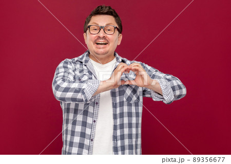 Portrait of happy handsome middle aged man in heckered shirt, eyeglasses standing with heart shape gesture hands and looking at camera with toothy smile. studio shot, isolated on dark red background 89356677