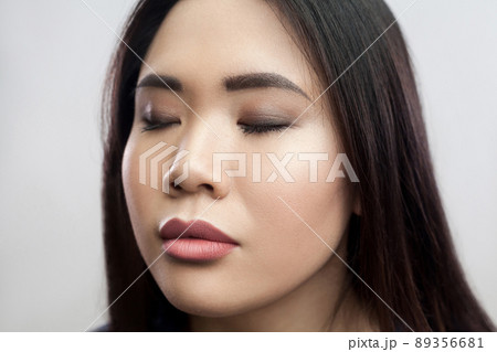 Closeup beauty portrait of calm beautiful brunette asian young woman with makeup, straight dark hair standing with closed eyes and relaxed face. indoor studio shot, isolated on light grey background. 89356681
