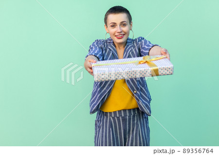 Portrait of friendly beautiful with short hair young woman in striped suit standing, giving you, congratulation you with present box, looking at camera. Indoor, isolated, studio shot, green background Portrait of friendly beautiful with short hair young woman in striped suit standing, giving you, congratulation you with present box, looking at camera. Indoor, isolated, studio shot, green background 89356764