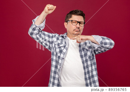 I am strong. Portrait of proud handsome middle aged business man in casual checkered shirt and eyeglasses standing and pointing at his biceps. indoor studio shot, isolated on dark red background. I am strong. Portrait of proud handsome middle aged business man in casual checkered shirt and eyeglasses standing and pointing at his biceps. indoor studio shot, isolated on dark red background. 89356765