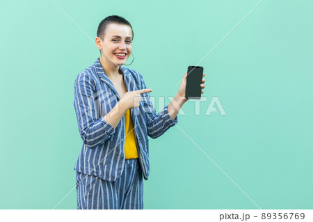Happy beautiful with short hair young woman in striped suit standing, holding phone and pointing finger to it, toothy smile and looking at camera. Indoor, isolated, studio shot, green background 89356769