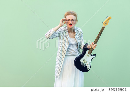 Portrait of young blonde woman in white shirt and striped blouse, electric guitar standing, holding eyeglasses and looking attentive at camera. indoor studio shot isolated on light green background. Portrait of young blonde woman in white shirt and striped blouse, electric guitar standing, holding eyeglasses and looking attentive at camera. indoor studio shot isolated on light green background. 89356901